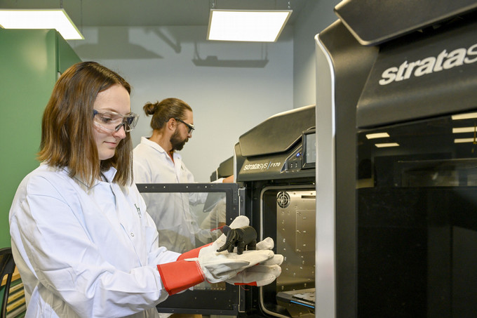 Two people in lab coats and safety gear work with a 3D printer, holding a small elephant figurine, in a bright lab.