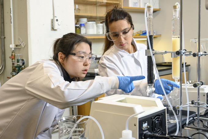 Two people in lab coats and safety goggles, wearing blue gloves, are closely examining and working with laboratory equipment