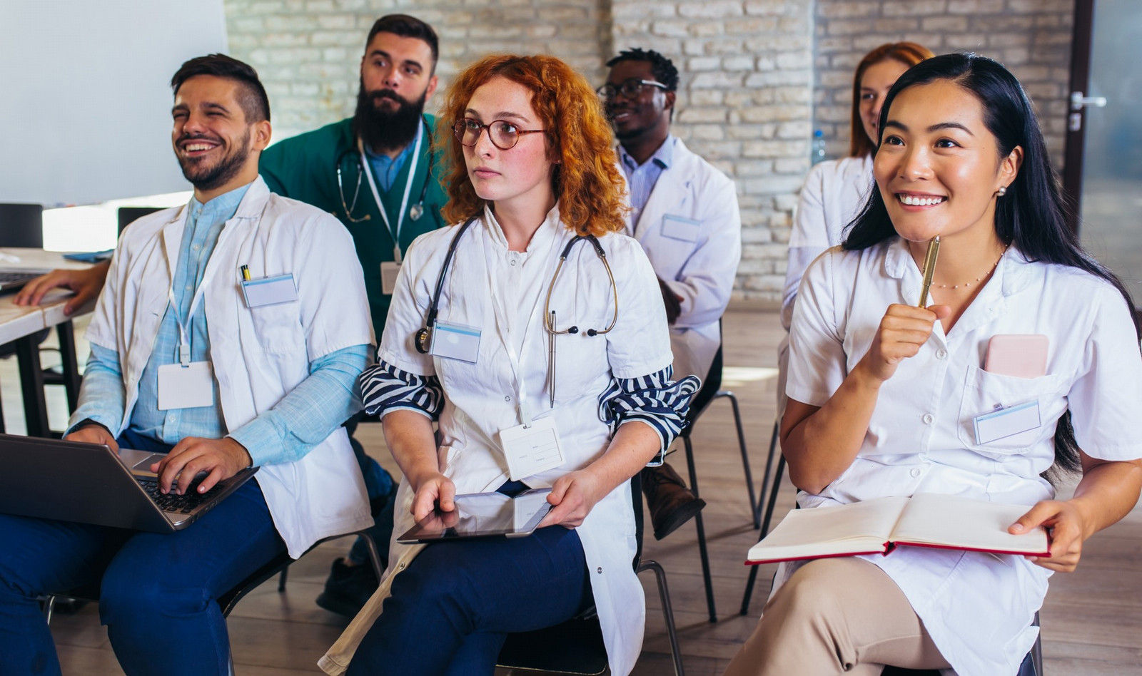 Group of diverse medical students attending a classroom lecture, wearing lab coats and taking notes with laptops and tablets.