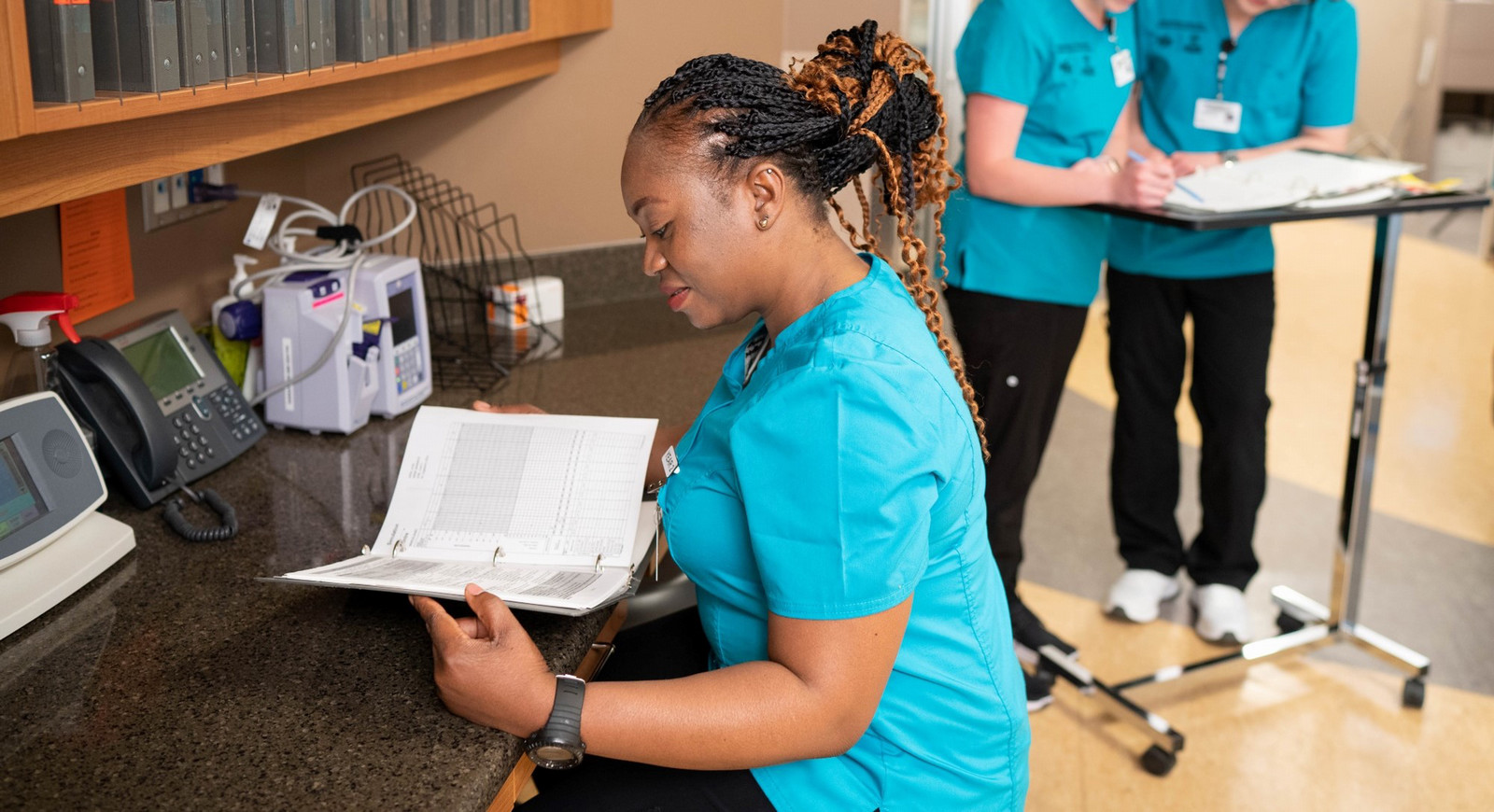 A nurse reading a book, with two people in the background talking
