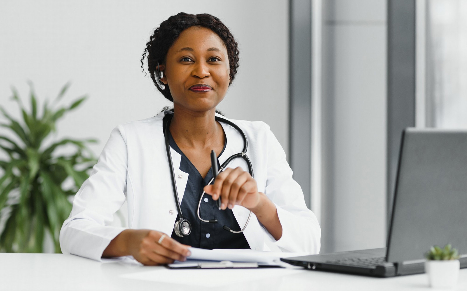 Smiling healthcare professional wearing a lab coat and headset, sitting at a desk with a laptop and medical notes in a modern office.