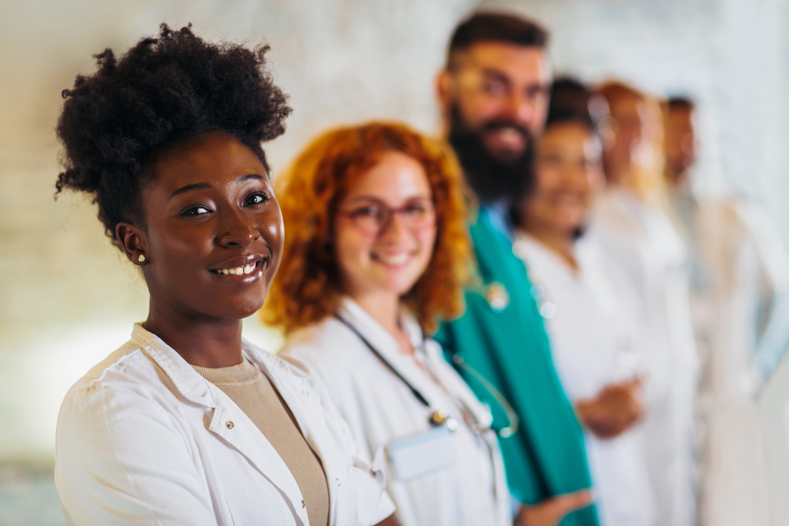 Diverse group of healthcare professionals smiling, wearing lab coats and medical uniforms, standing in a row in a clinical setting.