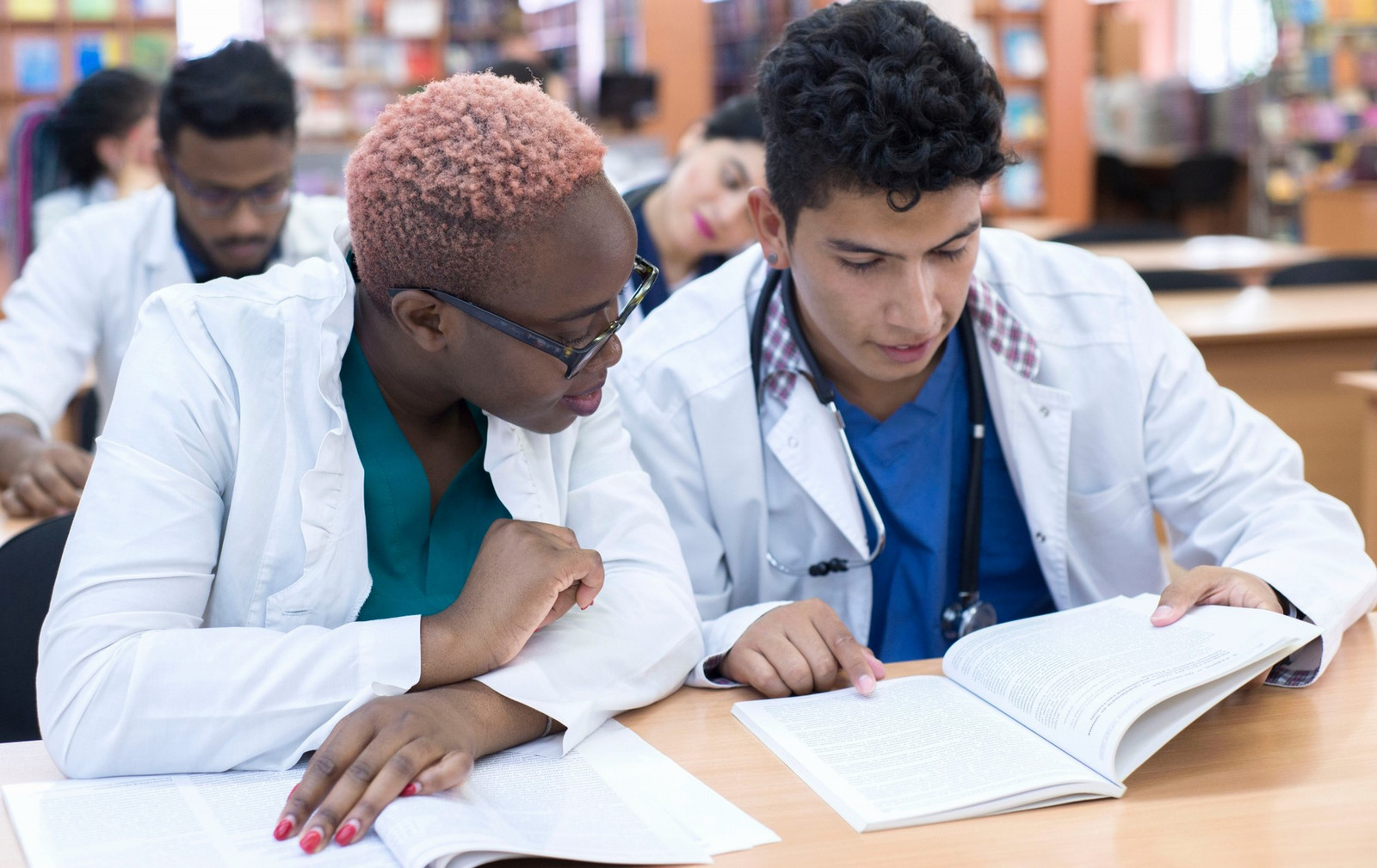 Medical students wearing lab coats studying together in a library, collaborating over textbooks in a healthcare education setting.