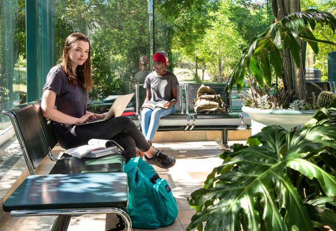 Student with a laptop posing for a photo with another student reading in the background of the atrium.
