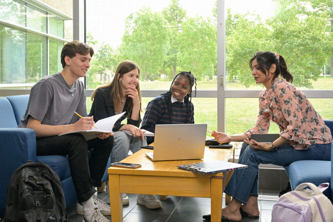 Five students sit in a bright lounge area, collaborating around a table with a laptop, notebooks, and backpacks, engaged in a friendly group discussion.