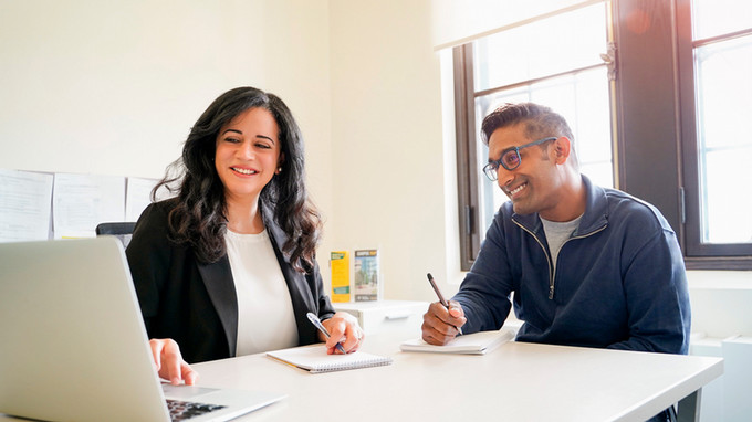 An academic advisor and student working together at a laptop, the student taking notes.