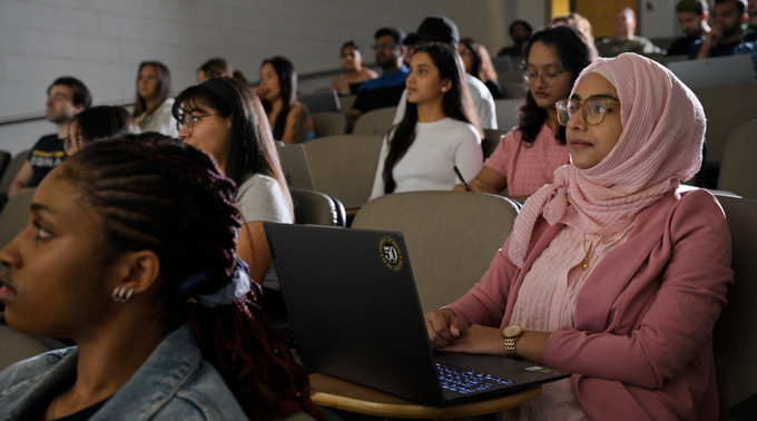 Students sit in a lecture hall, listening attentively, with one student using a laptop in the foreground.