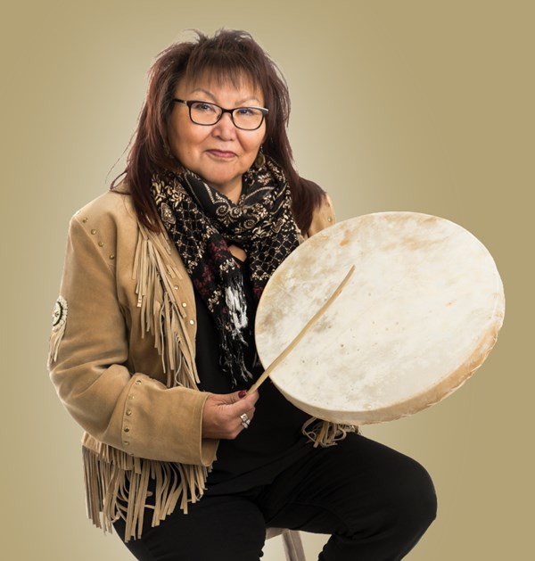 Photo of Rosalie Tsannie-Burseth holding a skin drum, wearing a sand-coloured jacket with fringe, patterened black scarf, black clothing and glasses.