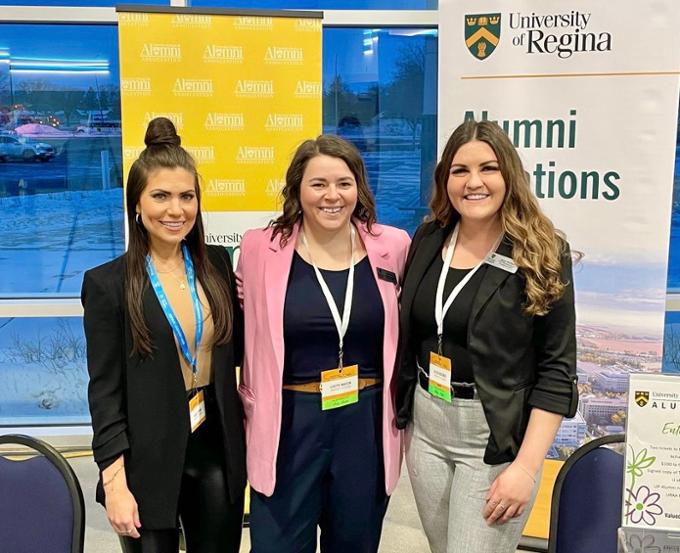 three women standing in front of banners for U of R alumni