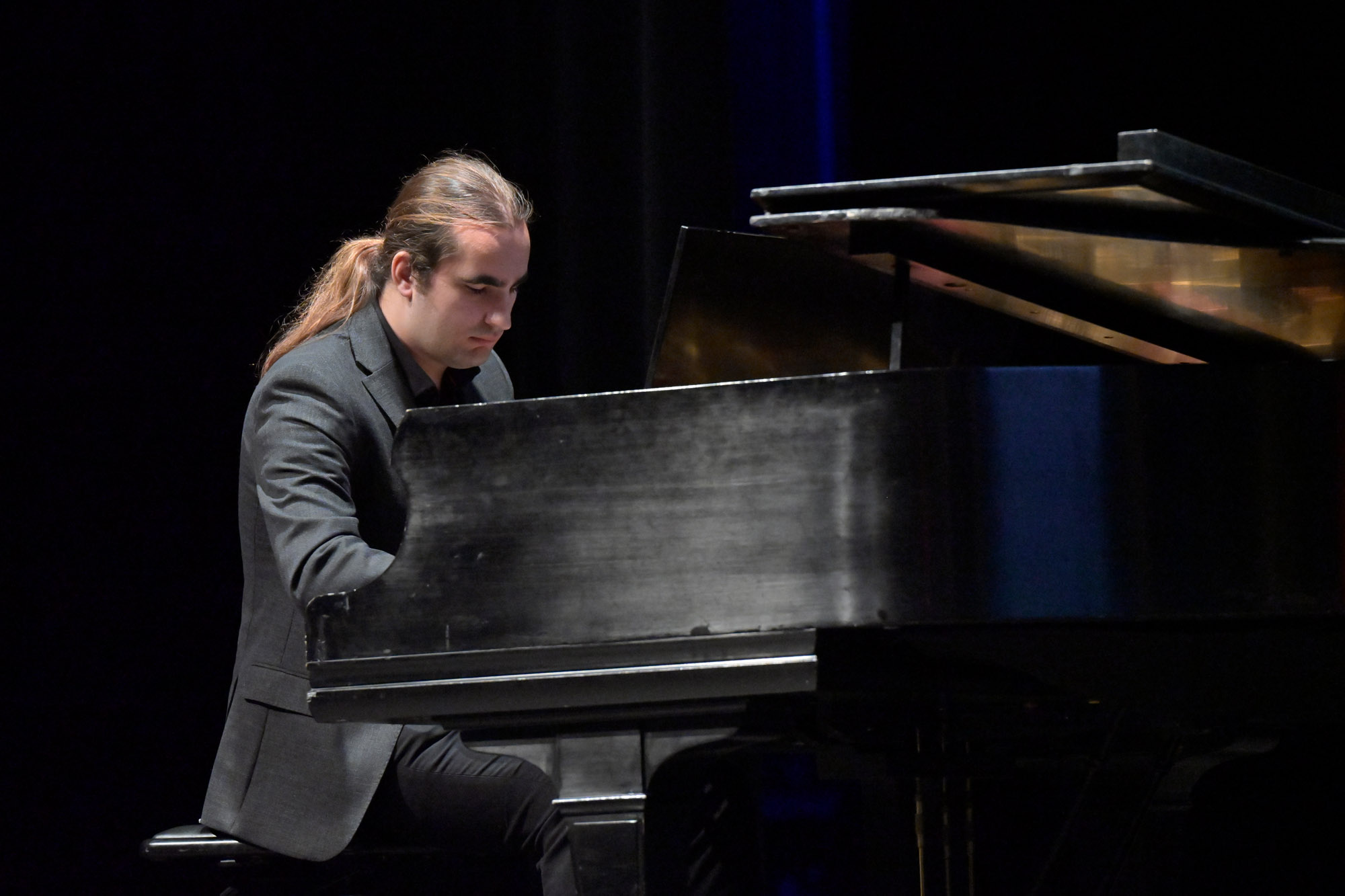 Man with ponytail wearing dark grey suit playing piano.