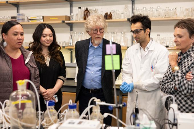 Four people listen to graduate student in lab