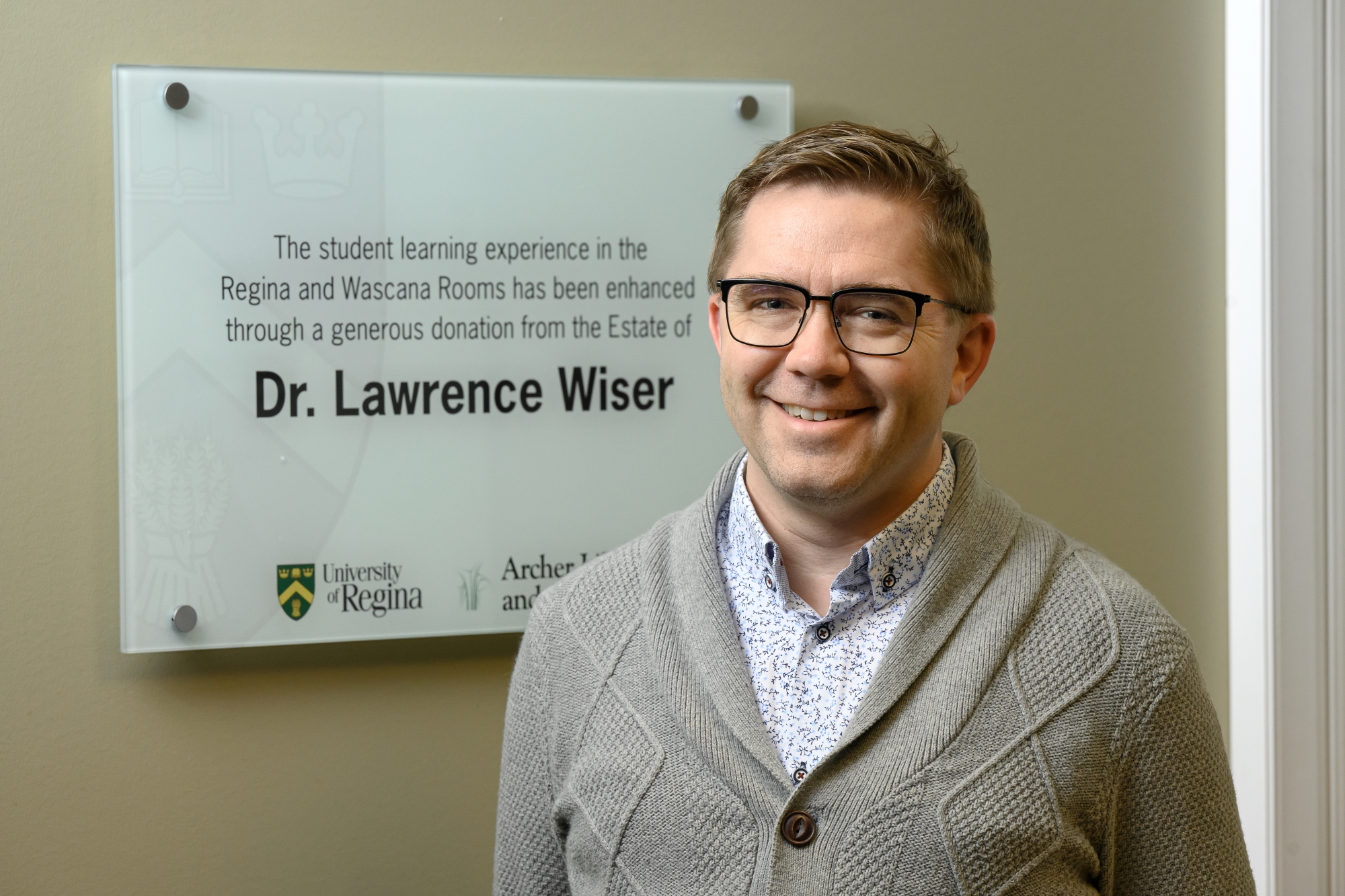 Dale Storie, Associate Dean (Research), stands next to the plaque honouring Dr. Lawrence Wiser in the Dr. John Archer Library and Archives.