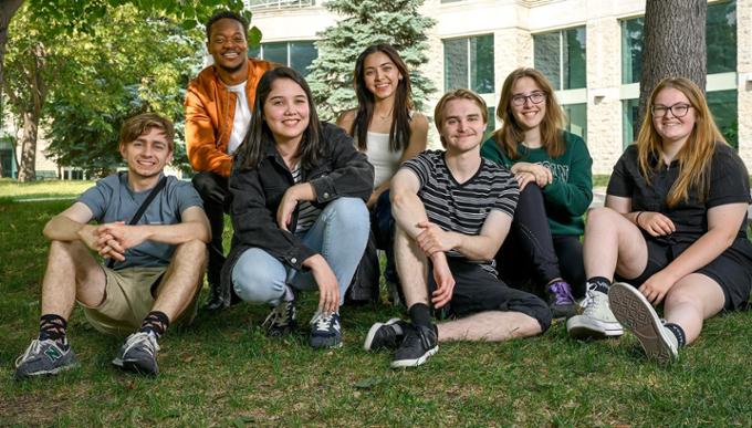 Group of students sitting outside on the lawn under trees looking at camera