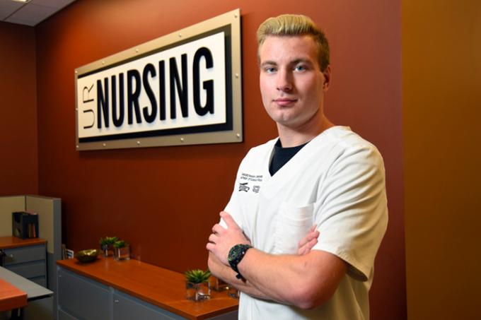 A student in scrubs stands in front of a Faculty of Nursing sign