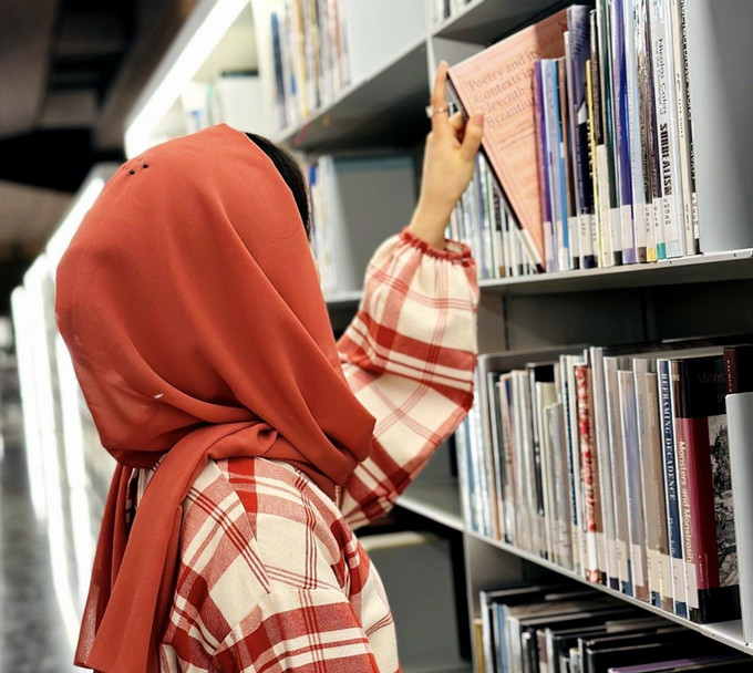 Woman in headscarf selects a book from a library bookshelf.