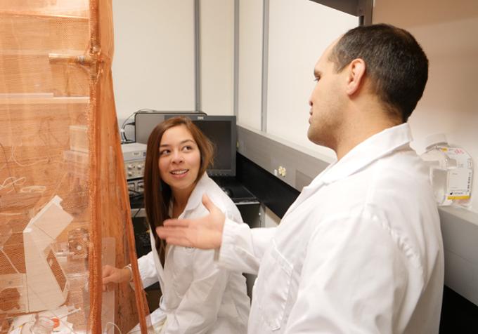 Two students in lab coats talk around a table with scientific equipment on it