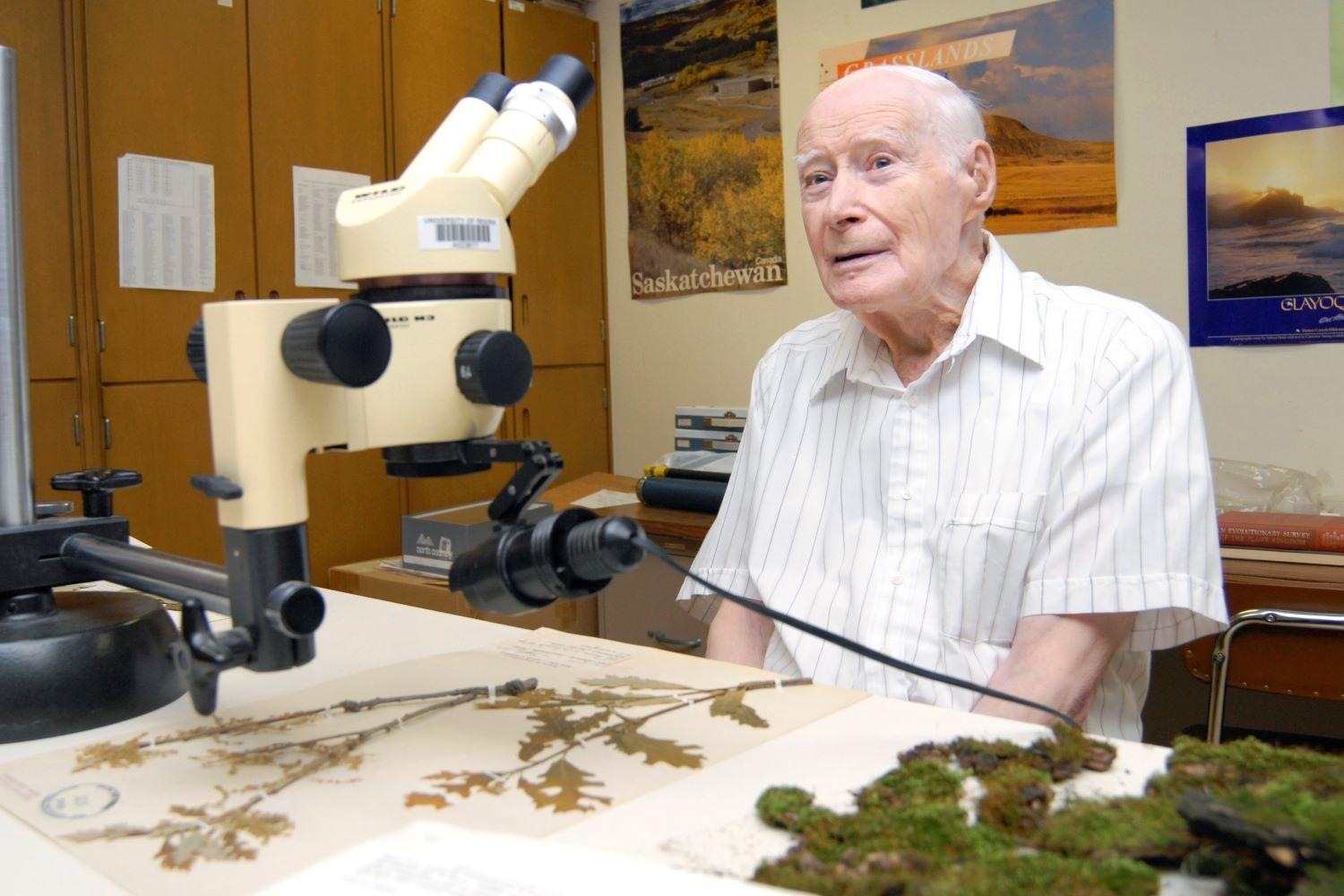 A man sits at a desk in front of a microscope and dried plant specimens.