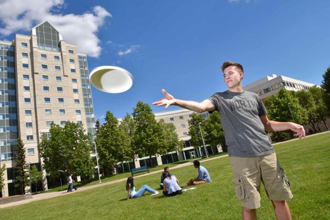 A student catches a frisbee in the quad area of campus on a sunny Summer day. Other students are seen in the background, sitting in grass relaxing and chatting.