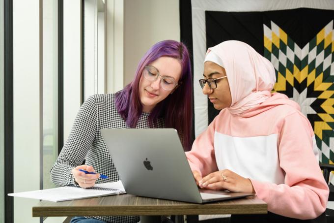 Two students sit in the study area of the Classroom Building lobby, laughing and chatting with one another.