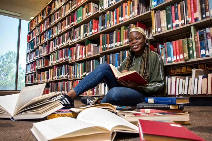 A smiling student sits on the floor of the library, looking at the camera and surrounded by books.