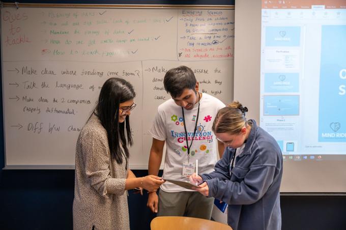 Team 1 discusses their idea in their classroom. The whiteboard behind them is full of notes.