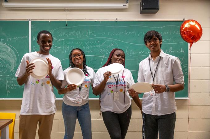 Team 6 poses for a picture with disposable plates. The chalkboard behind them has the name of their project written on it: SharePlate.