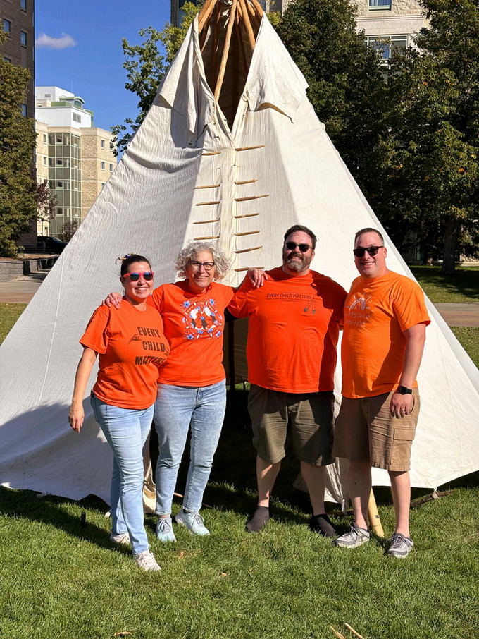Four smiling Faculty of Nursing team members wearing orange t-shirts standing in front of the tipi they just raised.
