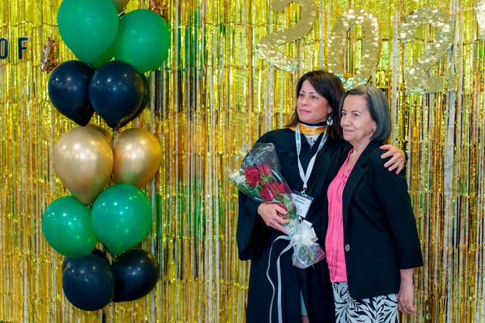female nursing graduate holding a bouquet of flowers has her arm around an older female both are standing in front of a gold tinseled back drop with green, black and gold balloons