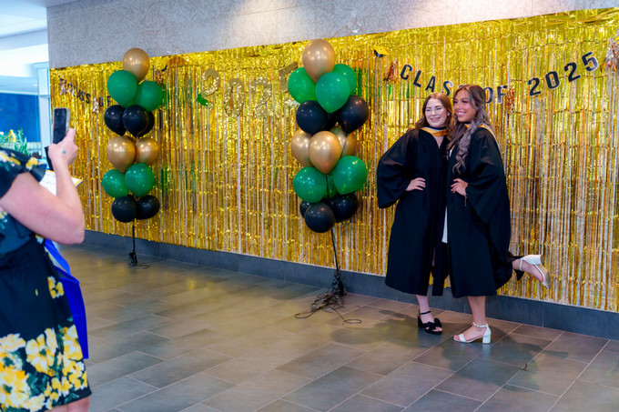 a candid photo of 2 female nursing grads in front of gold tinseled background that says class of 2025. 