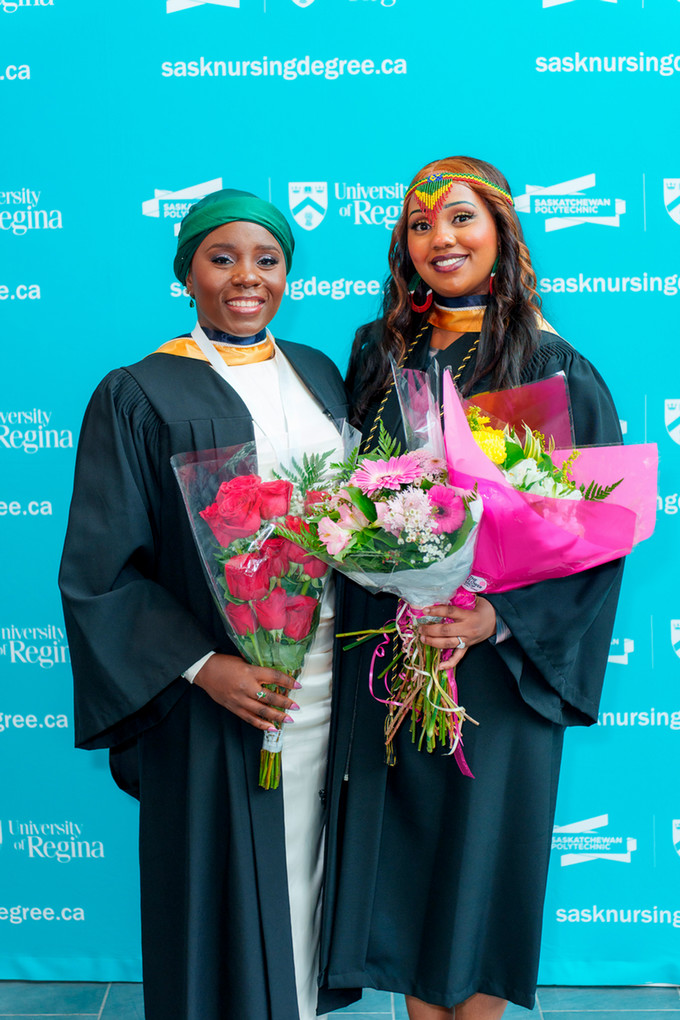 2 female bipoc nursing graduate holding bouquets of flowers. One is wearing an emerald green head wrap and the other a traditional beaded headband