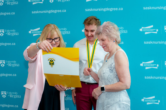 white male nursing grad winner of the deans medal pictured with both nursing deans looking at his diploma and smiling