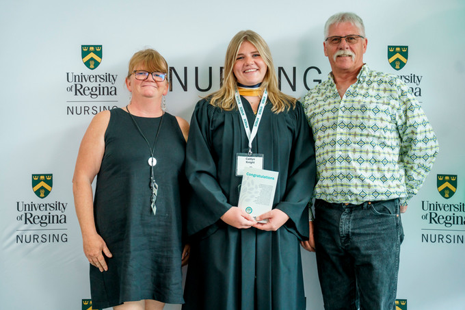 nursing graduate in black robe with gold ribbon holding her nursing pin surround on either side by her parents