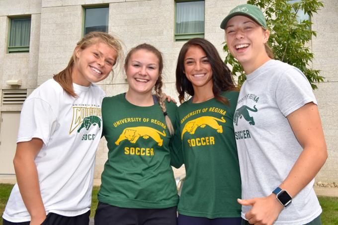 Four varsity athletes are posing for a photo outside on a summer day.