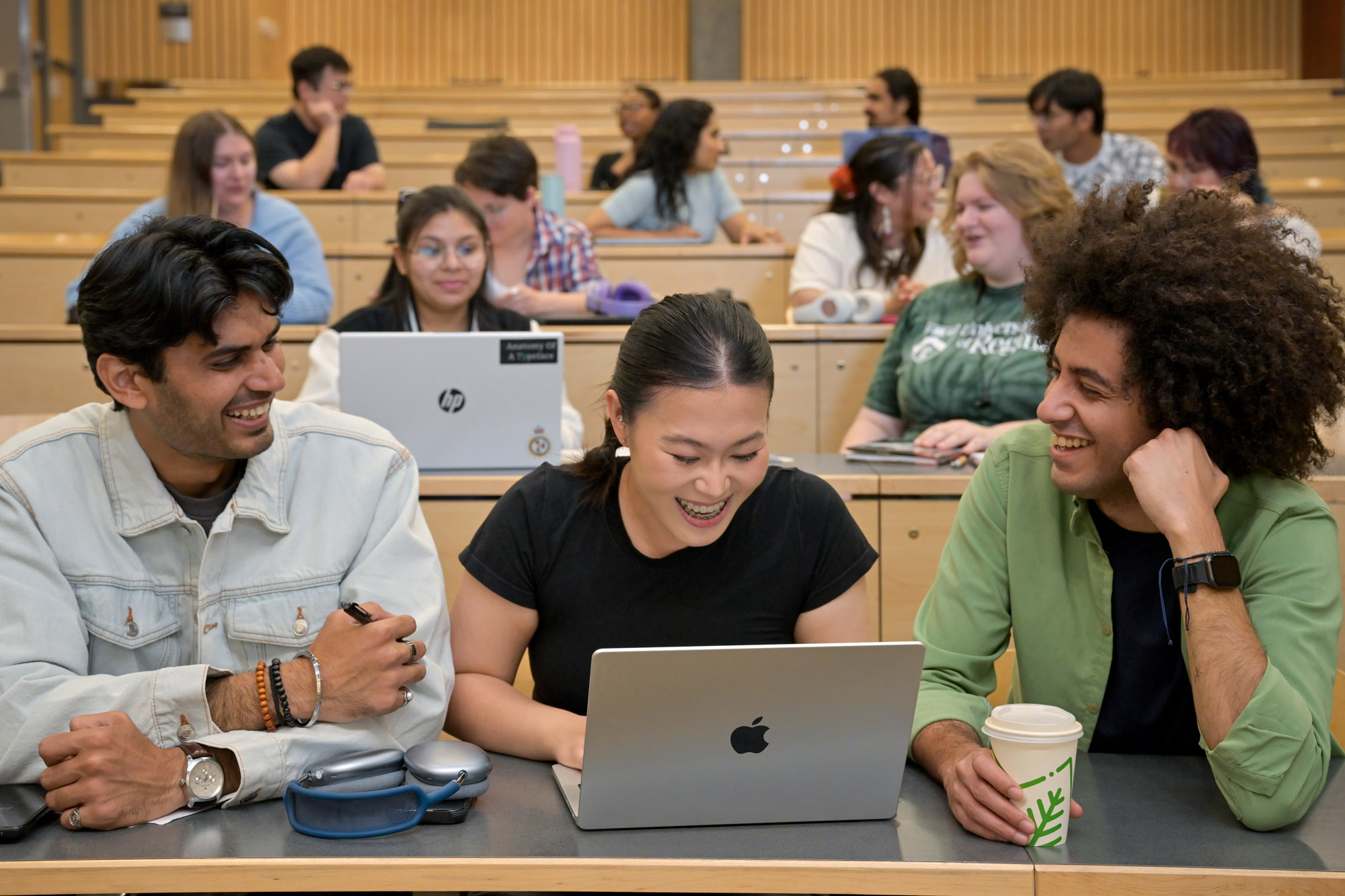 Three students sitting in lecture hall, looking at a computer, and smiling. 