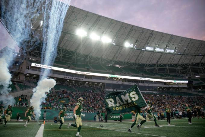 Regina Rams football players running onto field at Mosaic Stadium carrying a Rams flag. 