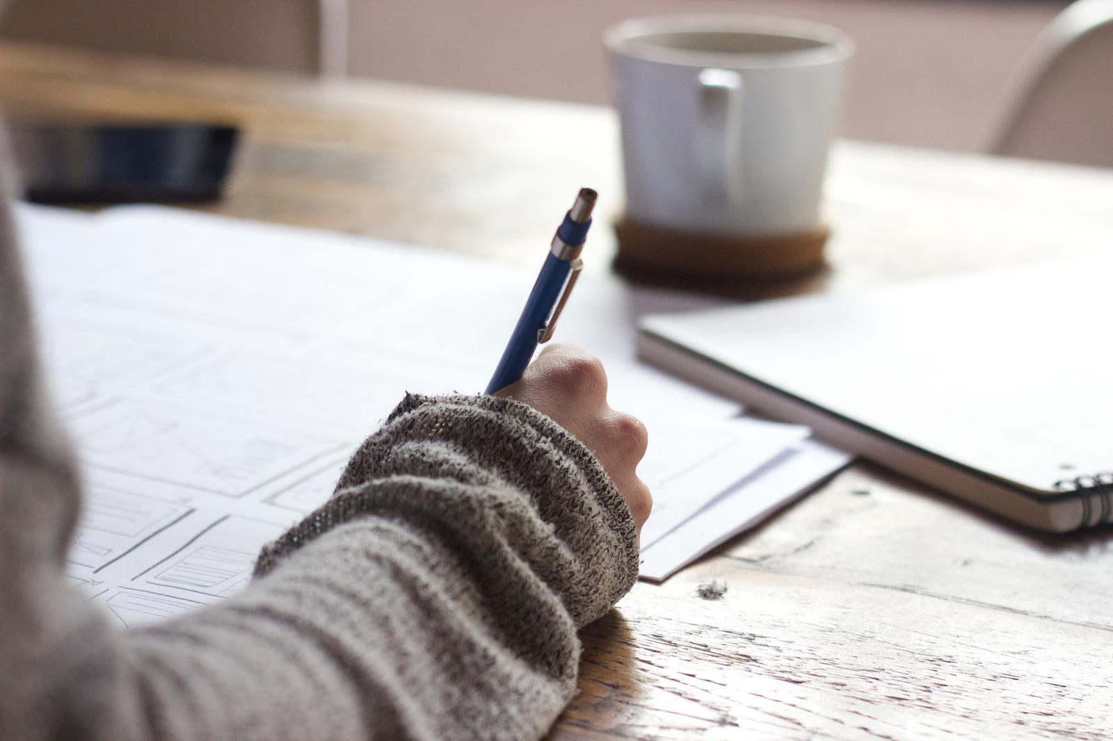 A student's hand with a pen is writing in a notepad. There is a coffee cup on the table as well.