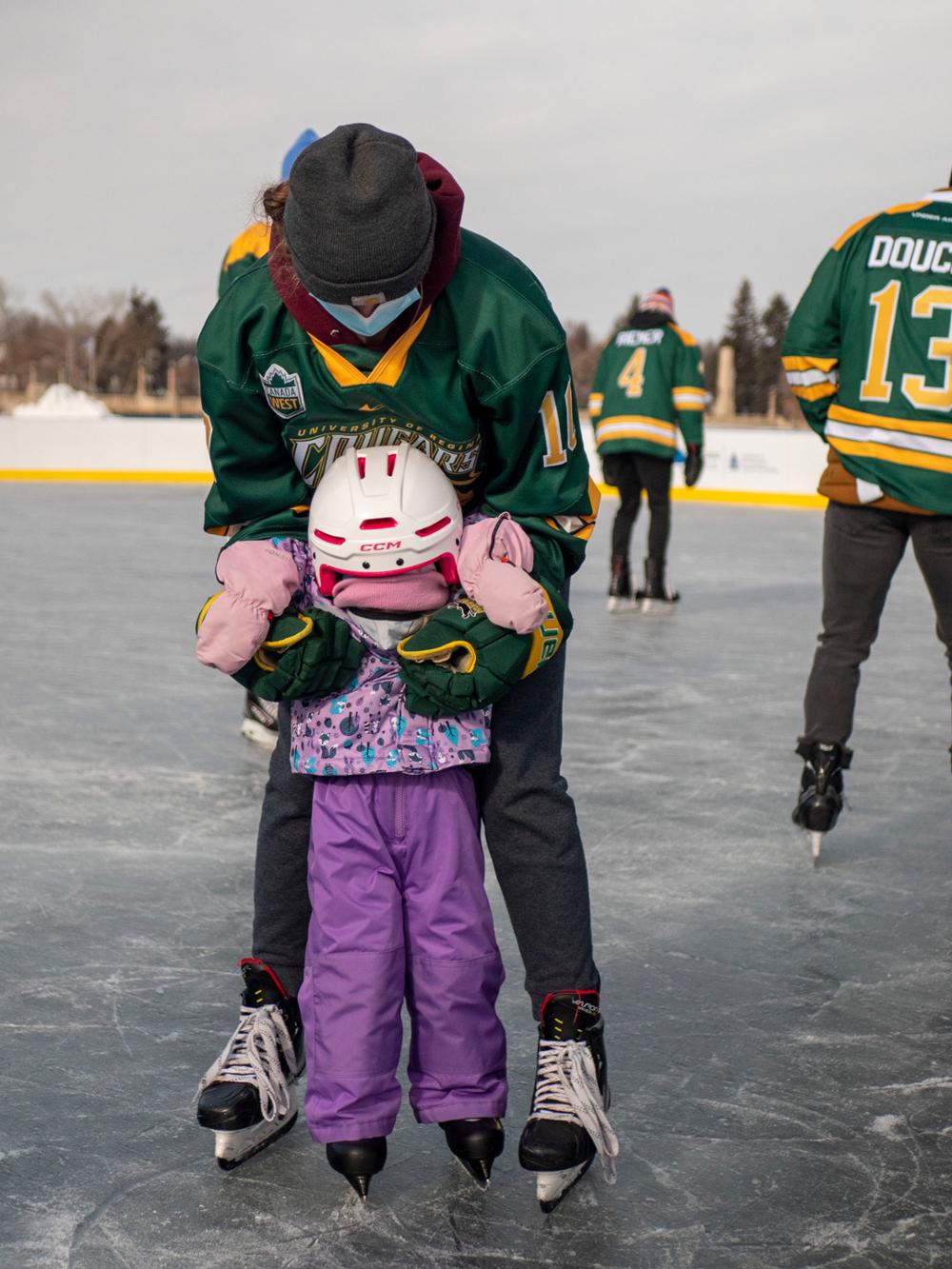 University student holding mittened hands of child while child to skate at outdoor rink.