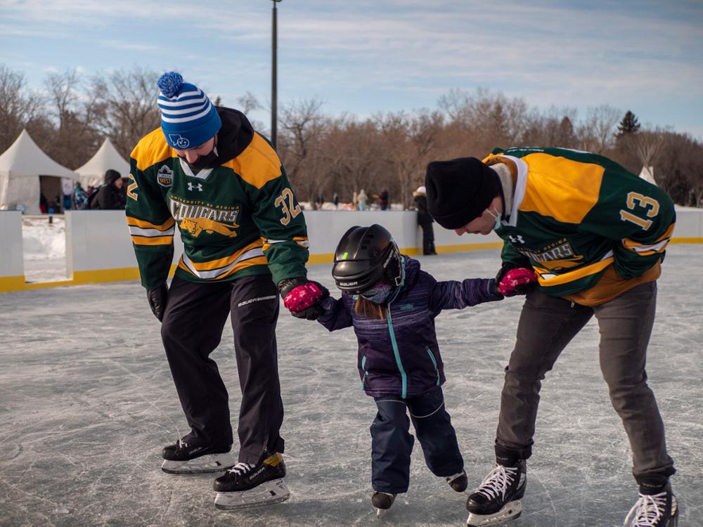 Two Cougar hockey players help a young child skate around.