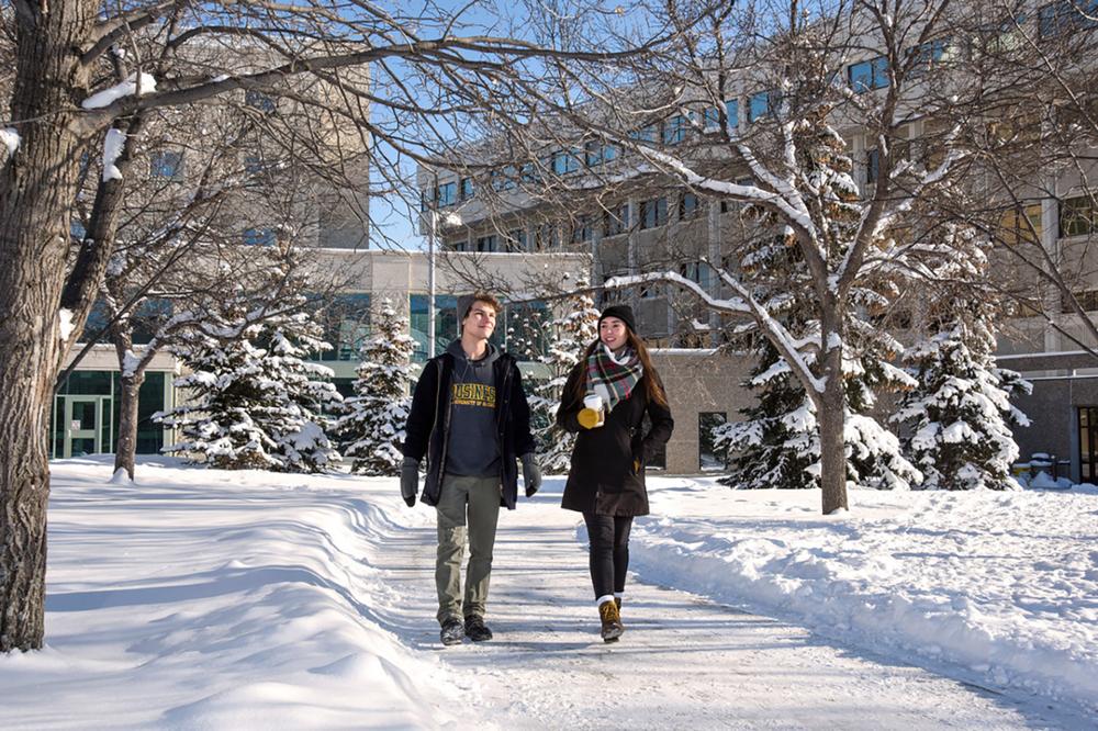 Two students walking side by side with snowbanks on either side of a campus sidewalk.