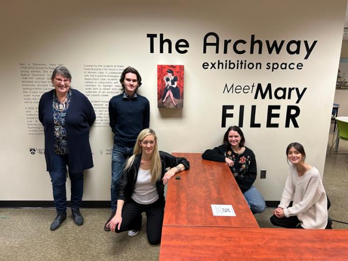 A professor and four students pose in front of entrance to art gallery featuring Mary Filer Exhibition.