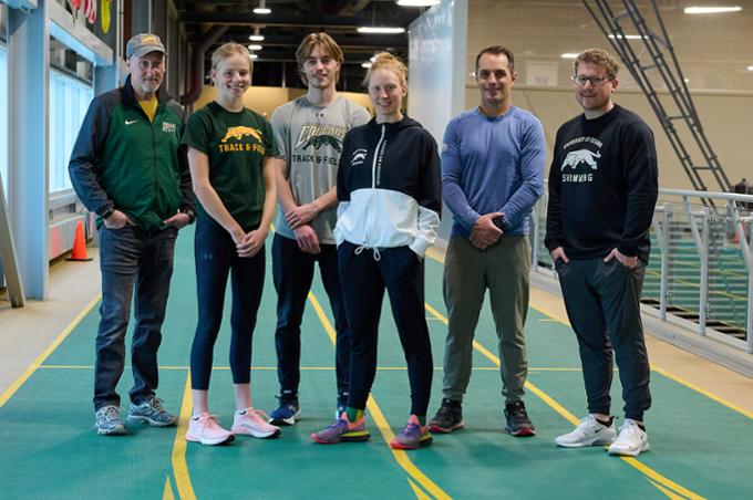 Three student-athletes stand on a track alongside three coaches.
