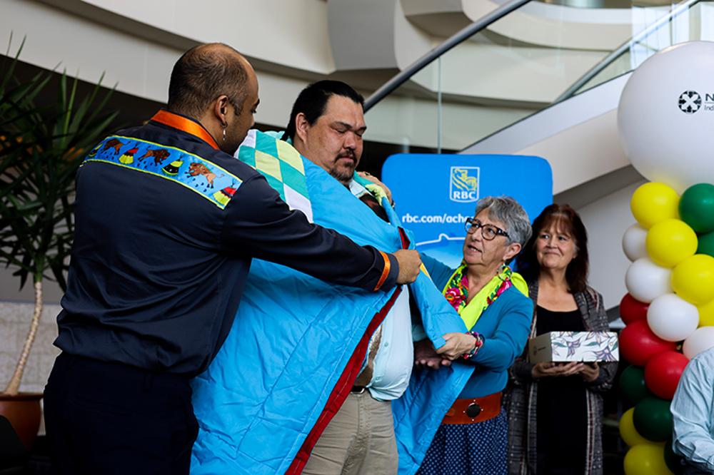 Two people wrap a student in a Star Blanket while a third person holding a gift box looks on.