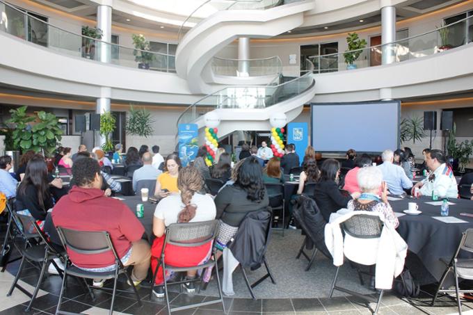 A room is filled with people sitting at round tables listening to a speaker who is sitting in front of a microphone