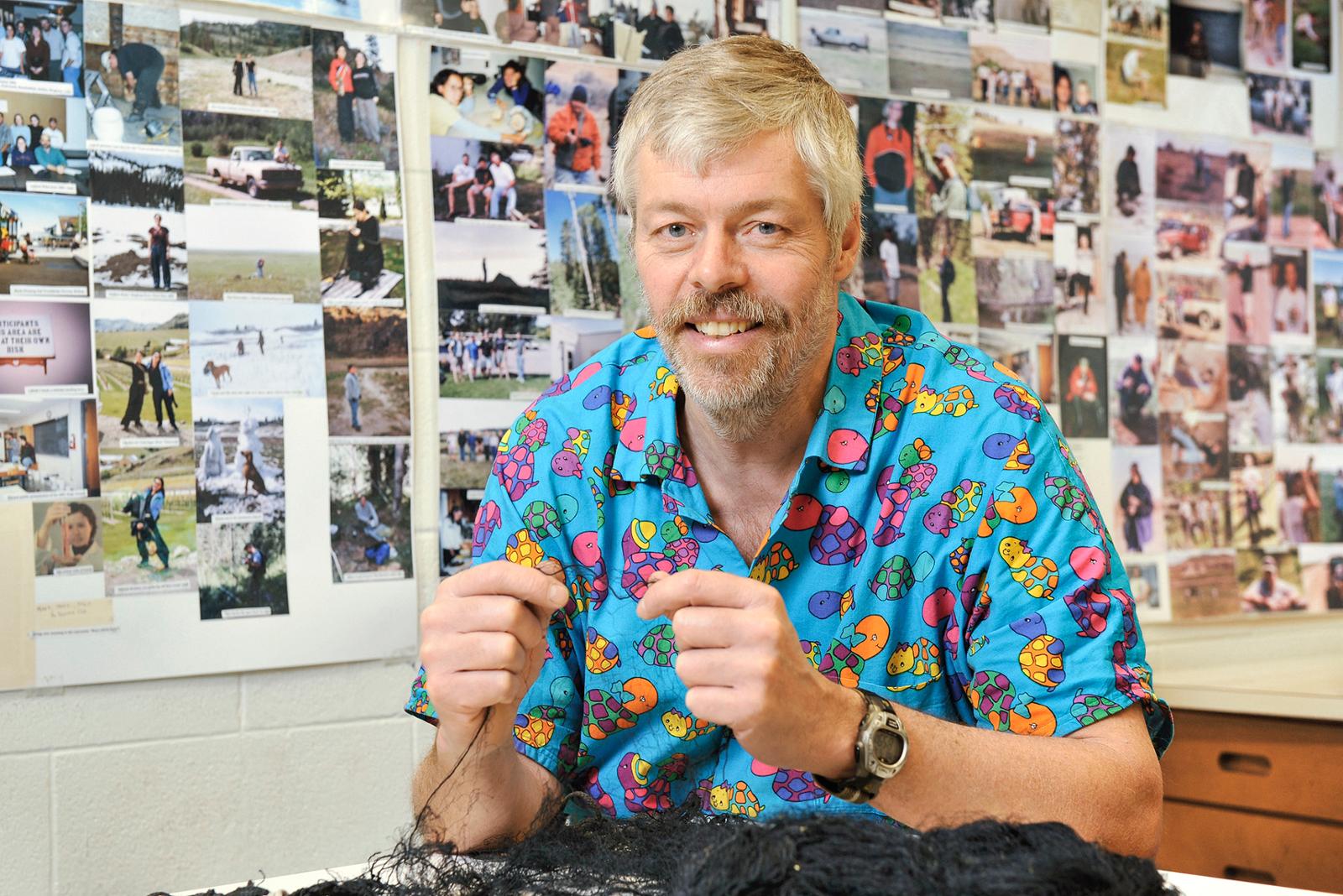 Biology professor Dr. Mark Brigham sitting at his desk with many photographs on wall behind him.