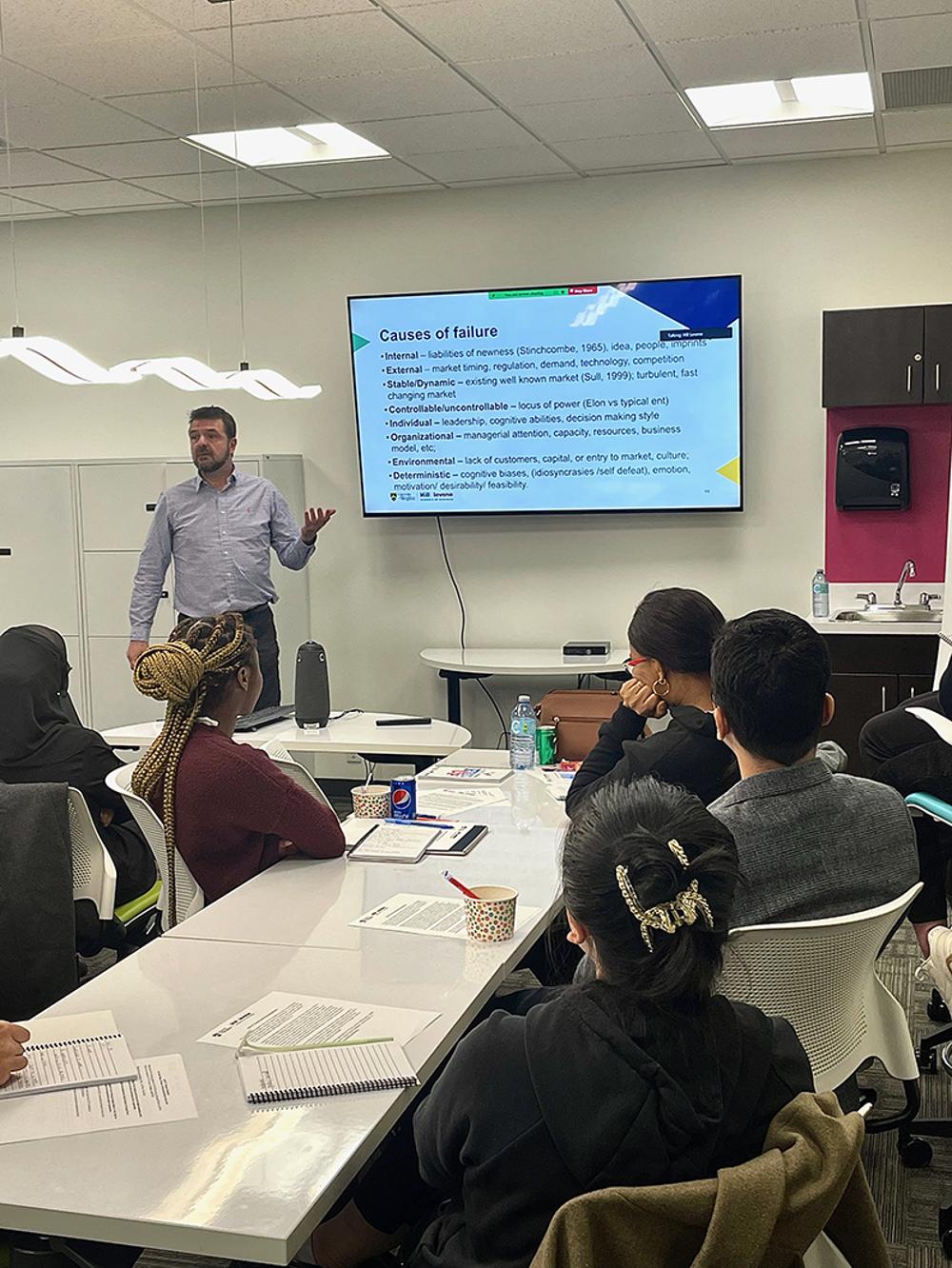 A professor talks to a room of students at long tables