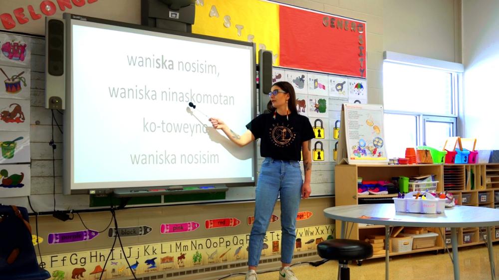 Indigenous teacher in a modern-day classroom points to Cree words on a whiteboard.