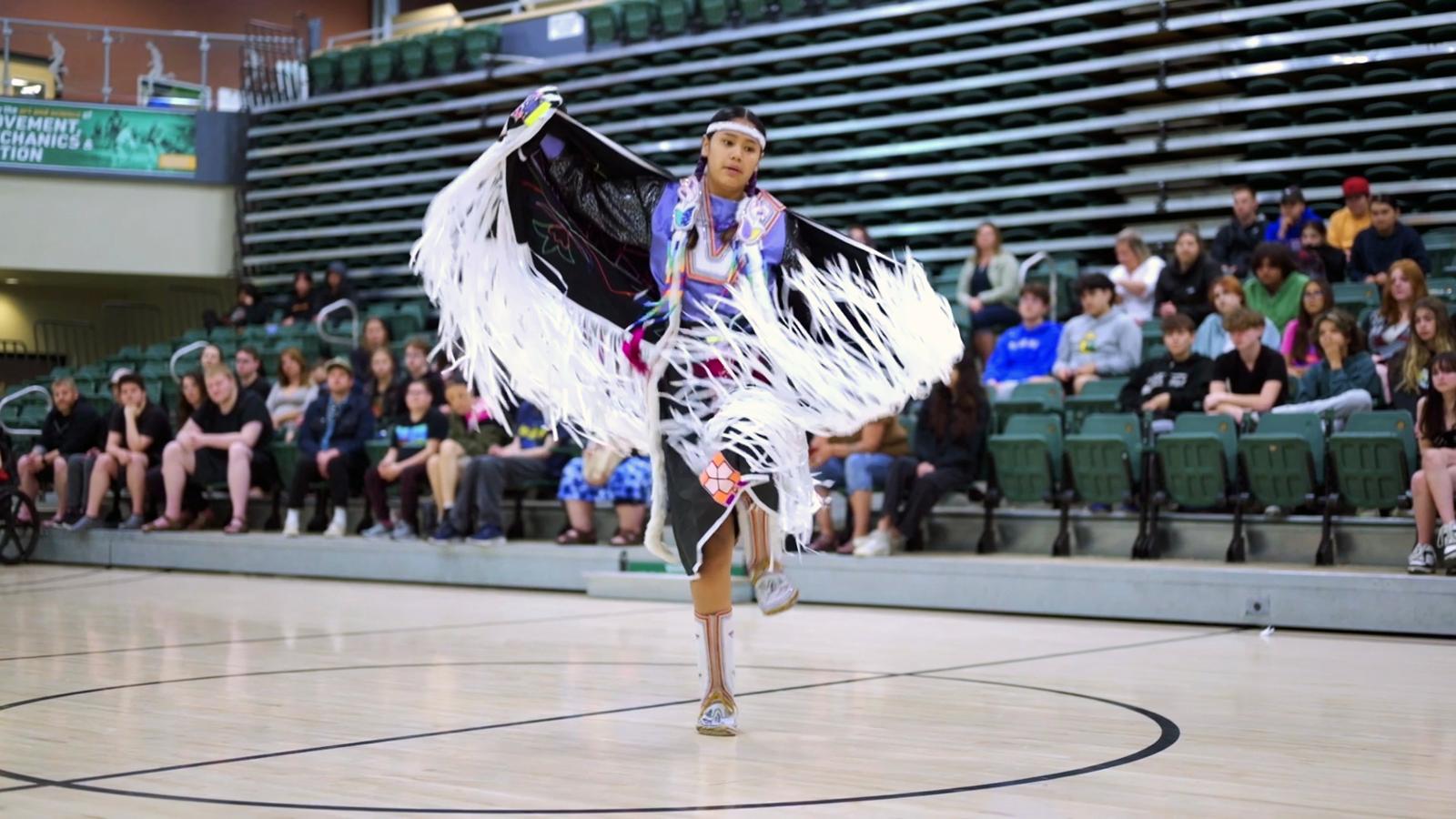 Young Indigenous woman shares Fancy Dress dance in front of crowd. 