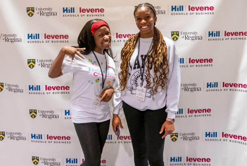 Two student-participants share an informal pose in front of the Hill and Levene Schools of Business banner