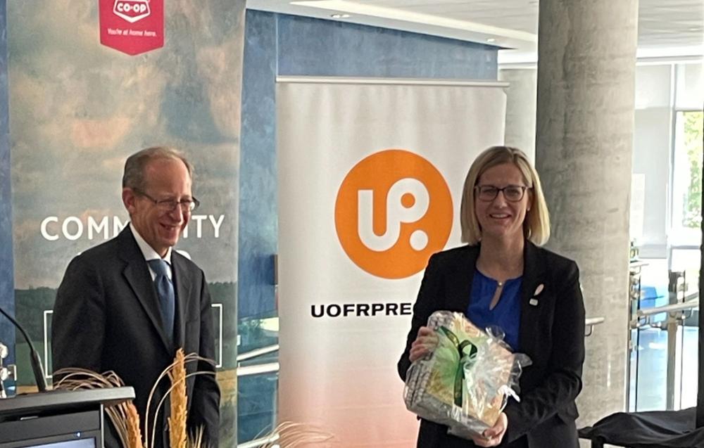 Man in suit stands next to woman in holding a gift-wrapped package of books from U of R Press. Pop-up banners from Federated Cooperatives Ltd and U of R Press are visible in background. 