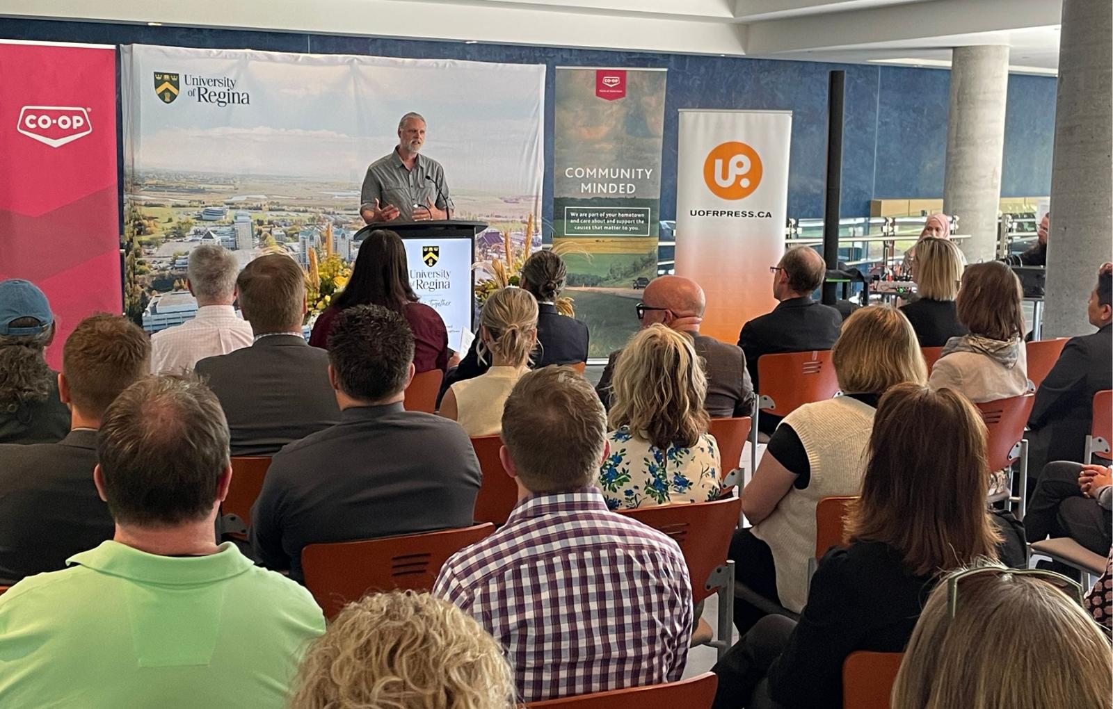 Man at podium addressing seated audience with banners visible in background branded for University of Regina and Federated Co-operatives Ltd.